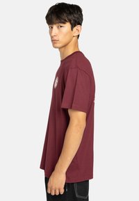 Camiseta de algodón burdeos con mangas cortas, cuello redondo y estampado de logo en el pecho y el costado, exhibida en una figura de pie.