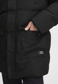 Svart pufferjakke med glidelås foran, sidelommer og logo-merke. Stoffet er glatt, og designet har en avslappet passform.