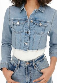 Mujer vistiendo una chaqueta de mezclilla azul clara cortada con botones de metal y jeans azul de cintura alta, con las manos en los bolsillos y una camisa blanca debajo.