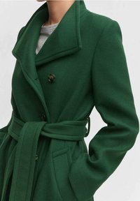 Cappotto verde scuro realizzato in tessuto testurizzato; presenta un colletto a scialle, doppi bottoni, una vita con cintura e tasche laterali.
