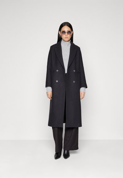 Lungo cappotto nero in lana con design doppiopetto, revers a punta, grandi bottoni e tasche laterali. Indossato sopra un dolcevita grigio e pantaloni a gamba larga.
