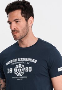 Navy blue cotton t-shirt with white graphic text "OMBRES MANSWEAR URBAN FASHION STYLE 1985" and small logo on the sleeve. Short sleeves, crew neck.