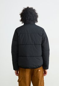 Geacă puffer neagră cu guler înalt, model cu pătrăți rectangulare și margine elastică. Purtați cu pantaloni din catifea călduroasă bej, cu buzunare laterale.