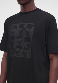 Άντρας που φοράει μαύρο T-shirt με σκούρο γραφικό τύπωμα εννέα ανδρικών προσώπων σε τρεις σειρές εμπρός.