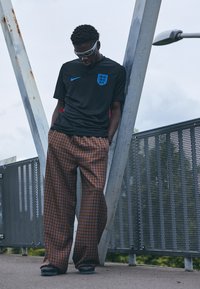 Juodas Nike marškinėliai su mėlynu Anglijos herbu, derinami su laisvai krentančiomis rudomis languotomis kelnėmis. Papuošta baltomis akinių nuo saulės.