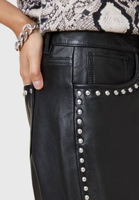 Zwarte leren rok met zilveren studs langs de zijkant, met een gladde textuur en zakken. De hand van het model rust op de tailleband.
