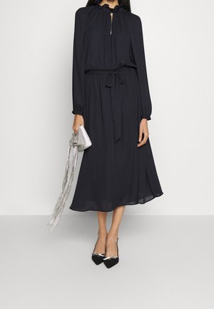 Robe bleu marine à manches longues avec col montant et ouverture en forme de goutte, taille cintrée, jupe fluide, accompagnée d’un sac à main argenté et de talons noirs.