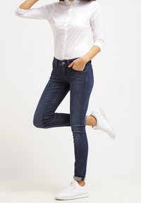 Jeans skinny azul oscuro hechos de denim, combinados con una camisa blanca ajustada con botones y zapatillas blancas. Diseño minimalista, costuras limpias.