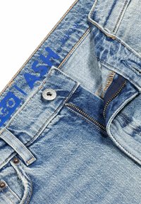 Jeans de mezclilla de color azul claro con una cremallera visible, remaches de cobre y texto azul impreso en el interior de la cinturilla. Presentan una textura desgastada.