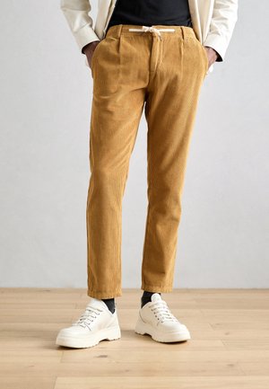 Mężczyzna ubrany w musztardowe sztruksowe spodnie z białym sznurkiem, czarne skarpety, białe trampki, czarną koszulę i kremową kurtkę stoi na drewnianej podłodze.