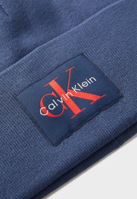 Berretto di maglina navy con una toppa rettangolare che presenta il testo rosso "CK" e "Calvin Klein" su uno sfondo blu scuro. Morbido al tatto, design piegato.