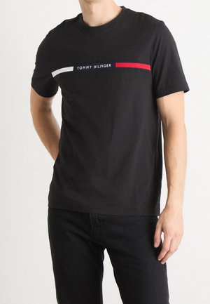 Mężczyzna w czarnej koszulce Tommy Hilfiger z białymi i czerwonymi poziomymi paskami na wysokości klatki piersiowej oraz w czarnych spodniach.