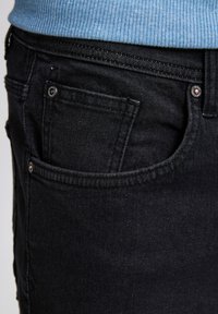 Zwarte denim jeans met een klassiek vijf-pocket design, stevige stiksels en een subtiele textuur. De tailleband is gedeeltelijk bedekt door een lichtblauwe trui.