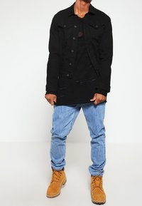 Veste en jean noire portée sur une chemise noire déchirée, associée à un jean bleu clair et des bottes de travail marron clair. Éléments de design simples et contemporains.