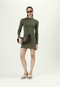 Vestido ajustado de manga longa verde-oliva, combinado com uma pequena mala preta de ombro e chinelos brancos, em um fundo claro e simplesmente.