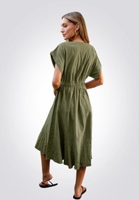 Vestido verde oliva de corte relajado, con mangas cortas y cinturilla elástica. Falda fluida con un acabado texturizado, combinada con sandalias negras.
