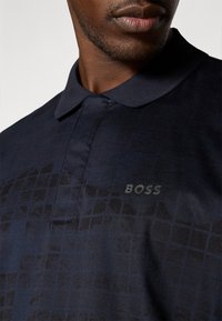 Jūras spēku polo krekls ar režģa rakstu, kuram ir apkakles apkakle un gaiši pelēks "BOSS" logo uz krūtīm. Gluda tekstūra.