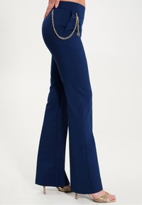 Žena v námořnických kalhotách se zvonovým střih a zlatým řetízkovým detailem, v béžových sandálech na vysokém podpatku, stojící na bílém pozadí.