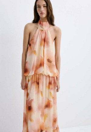 Jonge vrouw die een mouwloze halterjurk draagt met een zacht aquarelpatroon in oranje, perzik en beige, staand tegen een effen lichte achtergrond.