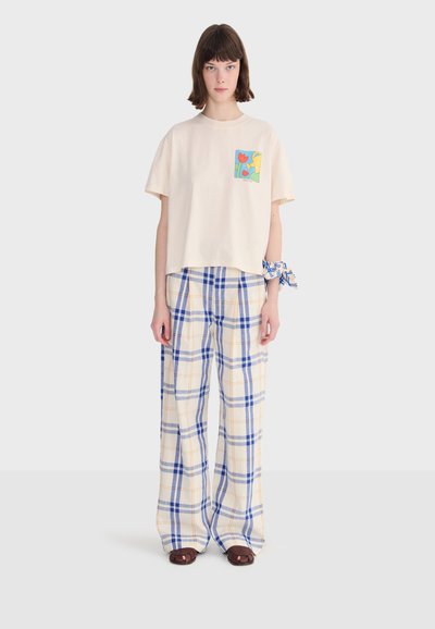 Femme portant un t-shirt crème avec une poche à imprimé floral coloré, un pantalon à jambes larges à carreaux bleu et beige, des sandales marron et un chouchou à motif bleu.