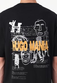 Svart bomull T-skjorte med iøynefallende gule "HUGO MANIA"-tekster, abstrakte hvite grafikker og håndskrevet stil på baksiden.