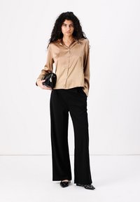 Satin beige blouse met knopen, zwarte wijde broek en zwarte clutch. Schoenen met zilveren accenten. Steil haar omlijst het gezicht.