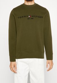 Uomo che indossa una felpa a maniche lunghe di Tommy Hilfiger di colore verde oliva con logo e pantaloni bianchi, mostrato dalle spalle ai fianchi.