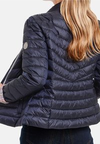 Marinblå quiltad pufferjacka med ståkrage, dragkedja fram och logotypdetalj. Har strukturerade paneler och en lätt design.