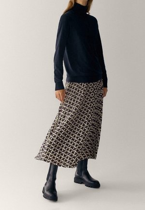 Personne portant un col roulé noir, une jupe midi à motifs et des bottes noires épaisses, debout devant un fond clair uni.