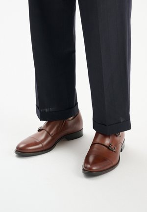 Brązowe skórzane buty typu double monk strap z zaokrąglonym noskiem, charakteryzujące się gładkim wykończeniem i czarną gumową podeszwą, w połączeniu z granatowymi spodniami w prążki.
