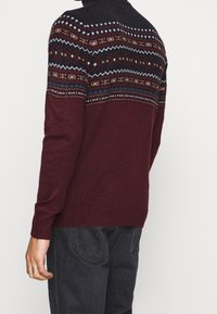 Mann in einem burgunderfarbenen Pullover mit mehrfarbigen nordischen Mustern auf dem oberen Rücken und den Schultern, kombiniert mit schwarzen Jeans.