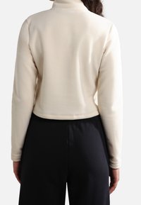 Chaqueta corta de forro polar crema con cuello alto, que presenta mangas largas y una textura suave. Combinada con pantalones negros de tiro alto.