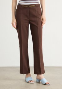 Pantaloni marroni su misura con pieghe verticali, caratterizzati da una cintura in catena dorata. Abbinati a mule aperti blu chiaro. Tessuto dal tocco liscio.