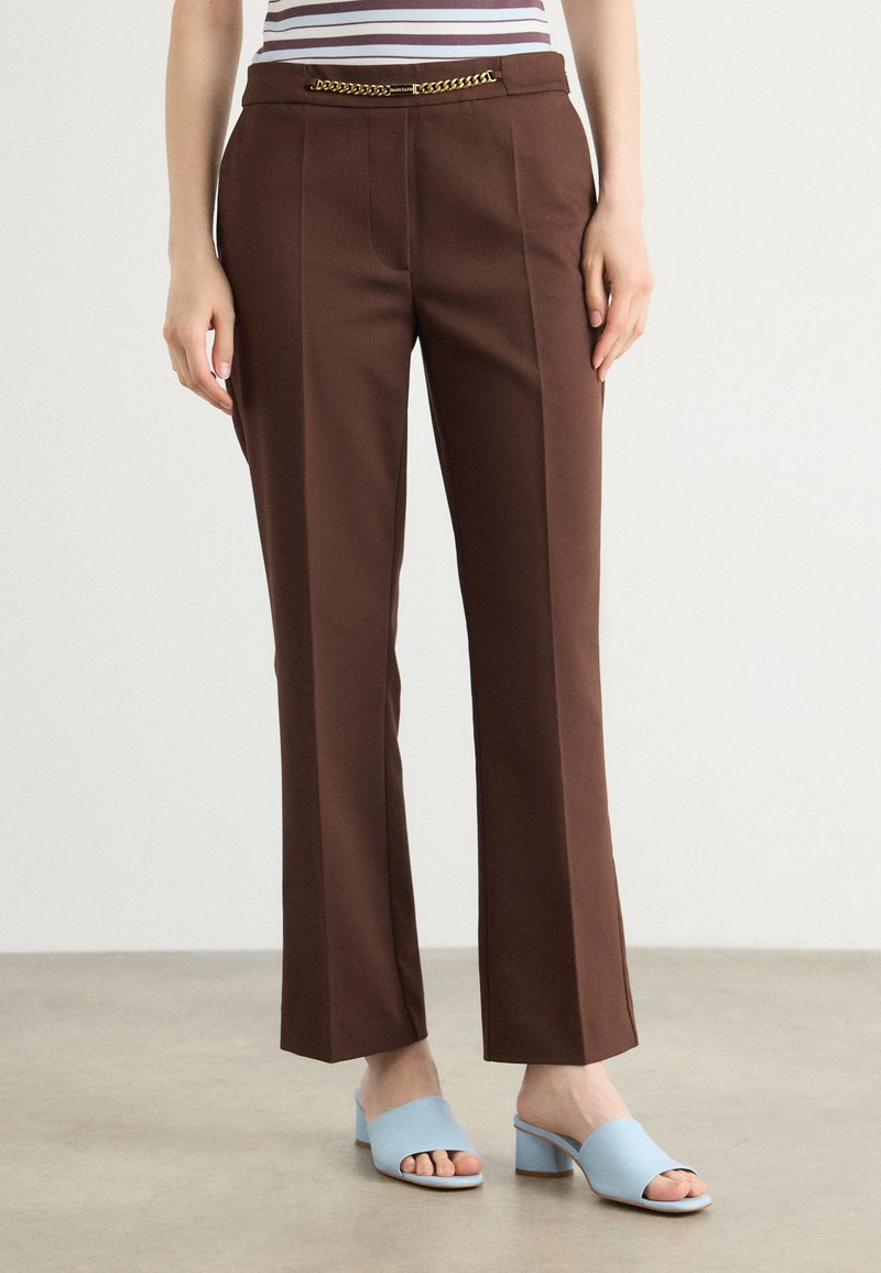 Pantaloni marroni su misura con pieghe verticali, caratterizzati da una cintura in catena dorata. Abbinati a mule aperti blu chiaro. Tessuto dal tocco liscio.