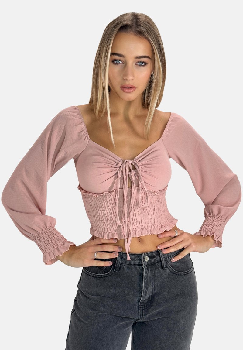Blusa corta rosa con lunghe maniche a sbuffo e vita smock. Presenta una chiusura frontale con legatura e una texture morbida. Abbinata a jeans a vita alta.