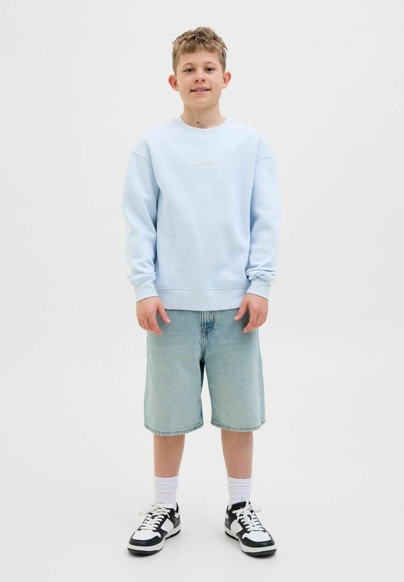 Felpa azzurra con collo rotondo, abbinata a pantaloni corti in denim chiaro fino al ginocchio e scarpe da ginnastica in bianco e nero, in piedi su uno sfondo semplice.