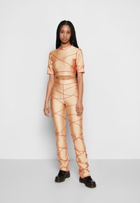 Conjunto bege de duas peças, com top cropped de mangas curtas e calças justas, apresentando detalhes em cruz vermelho, feito de um material liso e brilhante.