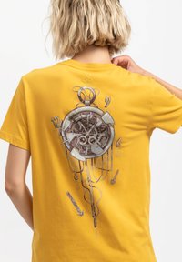 T-shirt di cotone gialla con una grafica dettagliata di un orologio da tasca con le lancette e i numeri, e linee stilizzate che si estendono verso il basso.