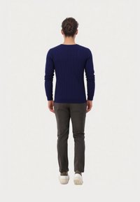 Persoon met bruin haar in een knot, gekleed in een geribbeld marineblauw tricot, donkere grijze broek en witte sneakers, staand met de rug naar de camera op een witte achtergrond.