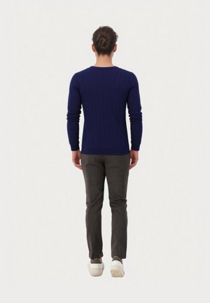 Persoon met bruin haar in een knot, gekleed in een geribbeld marineblauw tricot, donkere grijze broek en witte sneakers, staand met de rug naar de camera op een witte achtergrond.