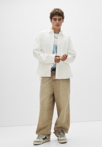 Weißes Hemd mit Knöpfen über einem hellblauen T-Shirt, kombiniert mit lockeren beigen Hosen und grauen Turnschuhen. Minimalistisches Design mit lässigem Schnitt.