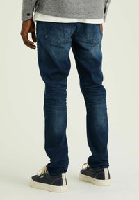 Szorosan illeszkedő farmernadrág sötétkék denim színben, enyhe kopással. Lapított hátsó zsebekkel és keskenyedő szárakkal rendelkezik. Sötét sportcipővel párosítva.