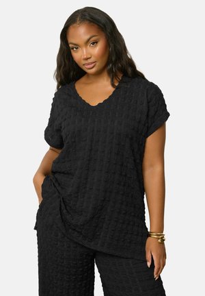 Kvinde med langt mørkt hår iført sort tekstureret top med korte ærmer og matchende bukser, accessoriseret med guldbøjler, poserer med hånden i lommen.