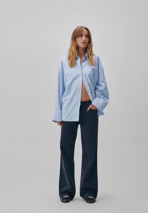 Camisa de botones de gran tamaño en azul claro con cuello en punta, combinada con jeans de pierna ancha negros y zapatos negros sin cordones.