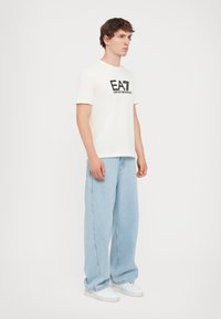 White short-sleeve t-shirt with black logo text on the front, paired with light blue loose-fit jeans and white sneakers.