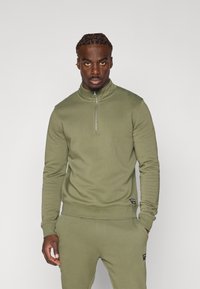 Felpa verde oliva con mezza zip, polsini e orlo a costine. Realizzata in un tessuto morbido con una texture liscia.
