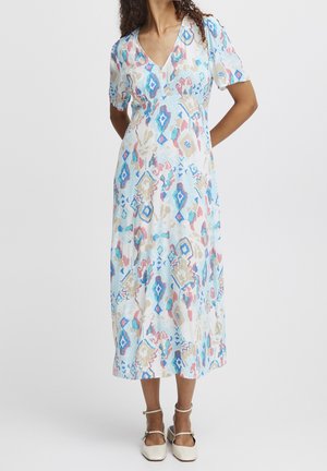 Femme portant une robe midi avec des motifs abstraits en bleu pastel, rose et beige et des chaussures blanches à bride cheville, debout, les mains dans le dos.