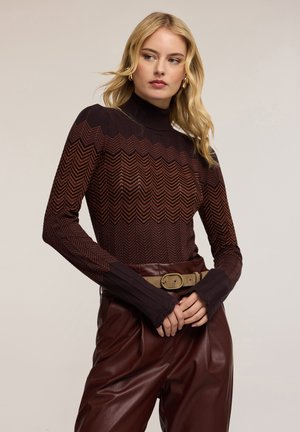 Suéter de cuello alto de punto con un patrón de chevron en tonos rojo oscuro y marrón, con mangas largas y puños acanalados, combinado con pantalones de cintura alta.