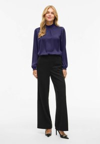 Lilla satengbluse med høy hals og rynkedetalj, kombinert med svarte vide bukser og svarte spisse hæler med gullaccent.