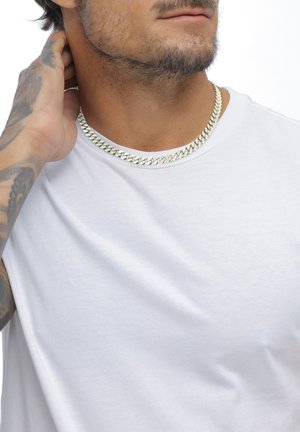 Mann trägt ein weißes T-Shirt und eine silberne Gliederkette, mit tätowiertem Arm, der hinter seinem Nacken greift, vor einem schlichten weißen Hintergrund.